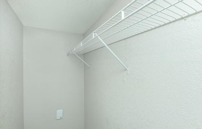 Empty closet corner with light gray walls and a white wire shelving unit mounted on the wall. The shelving is positioned at the top, with one side secured to the wall, and there is a white power outlet visible on the wall below the shelving. The space appears clean and uncluttered.