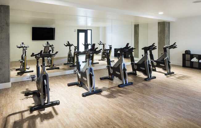 A room with a row of stationary bikes and a television mounted on the wall.