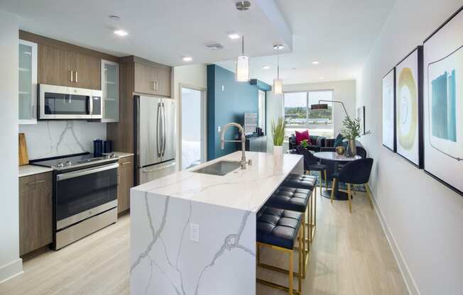 Woodland Hills, CA Apartments - The Q Variel - Kitchen with Luxury Vinyl Flooring, Quartz Waterfall Kitchen Island, Advanced Wash Dishwasher, LED Energy-Efficient Lighting Fixtures, and Open-Concept Layout with the Dining Room and Living Room which have High Ceilings and Expansive Windowsirs