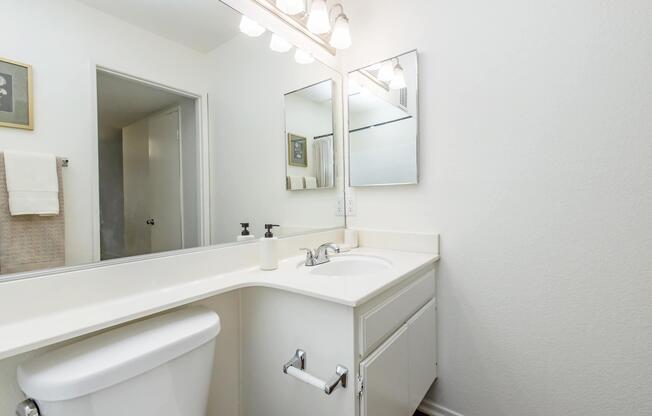 A clean, modern bathroom featuring a white sink and countertop, a large mirror with light fixtures above, and a towel hanging on the wall. The walls are painted in a light color, and the floor has dark wood accents. A minimalistic and uncluttered aesthetic is evident throughout the space.