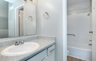 A clean, modern bathroom featuring a single sink countertop with a mirror above, a towel ring, and a bathtub with a shower curtain. The walls are painted a light color, and the floor has a wood-like finish, creating a bright and airy atmosphere.