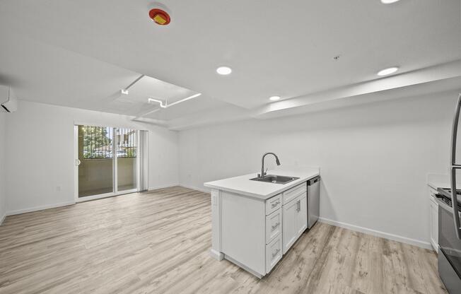 Bright, modern open-concept kitchen and living area with white cabinetry, a stainless steel sink, and appliances. The space features light wood flooring, recessed lighting, and a sliding glass door leading to an outdoor area. The walls are painted in a neutral tone, creating a spacious and inviting atmosphere.