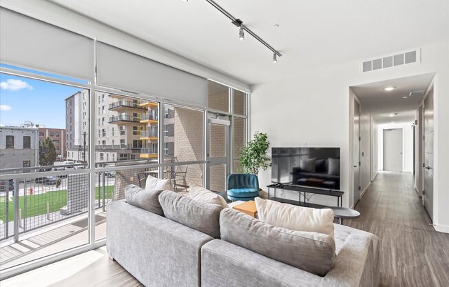 A living room with a grey couch and a balcony with a view of the city.