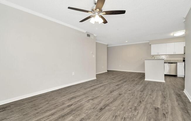 Modern interior of a spacious living area featuring light-colored walls, a ceiling fan, and wood-style flooring. The open layout includes a kitchen with white cabinets and appliances visible in the background, creating a bright and inviting atmosphere.