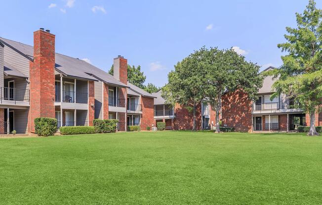 A well-maintained apartment complex featuring two-story buildings with brick exteriors. The lawns are lush and green, with trees providing shade. Clear blue sky and a serene atmosphere create a pleasant residential environment.