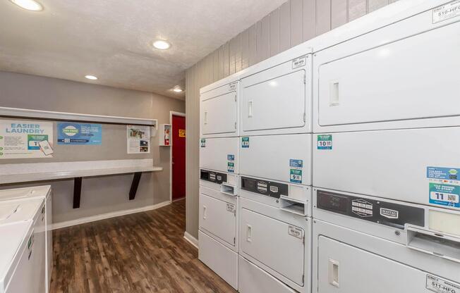 A well-lit laundry room featuring multiple stacked washing machines and dryers against a light gray wall. There is a countertop area with laundry supplies nearby, and informational signage on the walls. The flooring is wooden, adding a cozy feel to the space.