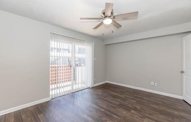A spacious, light-filled room with a ceiling fan and laminate flooring. The wall is painted in a soft gray color, and a large sliding door with vertical blinds leads to an outdoor area, enhancing the room's brightness and openness.