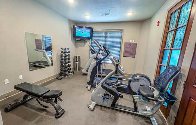 Compact fitness center at The Dorset Apartments featuring cardio machines, weight bench, dumbbells, and a mounted TV.