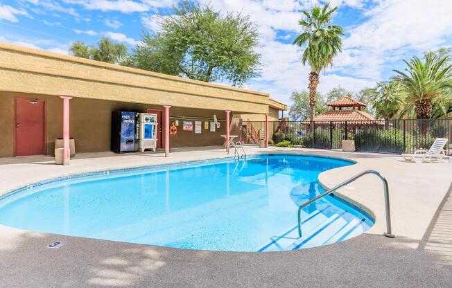 A serene outdoor swimming pool with a clear blue surface, surrounded by palm trees and a gated area. Lounge chairs are positioned nearby, and a vending machine is visible. The sky above is partly cloudy, adding to the tranquil atmosphere.