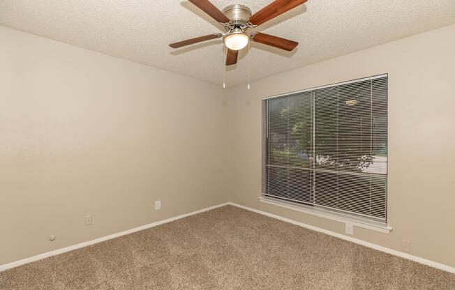 A small, empty room with beige walls and plush carpet. The ceiling features a modern ceiling fan with wooden blades. A large window with blinds allows natural light to enter, looking out to greenery. The space is unfurnished, ideal for personalization.