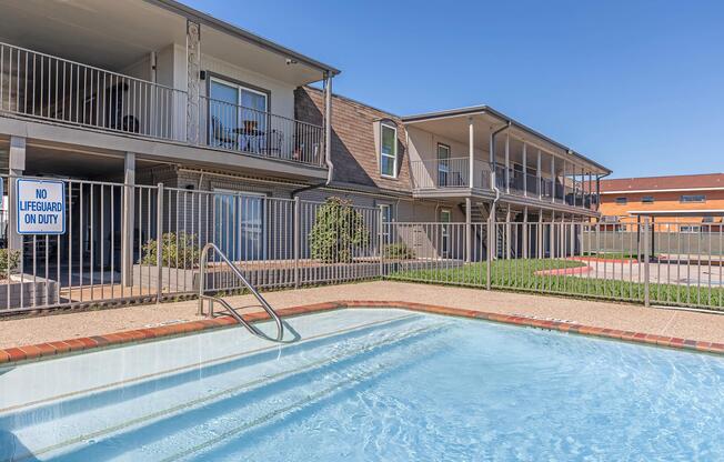 A clear swimming pool surrounded by a fence, with a "No Lifeguard on Duty" sign visible. In the background, there are two-story residential buildings with balconies. The area is well-lit and features green grass and walkways. The sky above is bright blue, indicating clear weather.