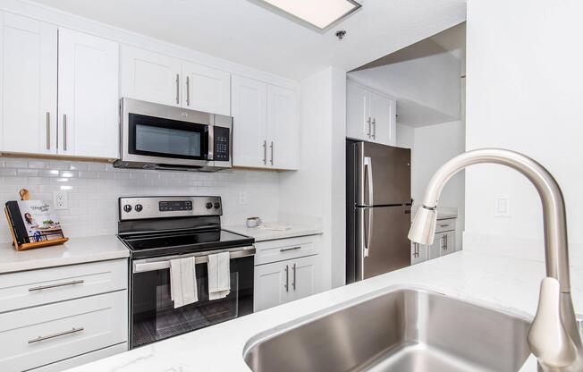 A modern kitchen featuring white cabinetry, a stainless steel microwave, and an oven. There is a double-basin sink with a stylish faucet in the foreground, and a refrigerator in the background. The countertops are made of a light-colored stone, and there's a small decorative item on the countertop.
