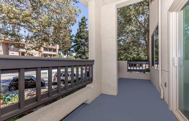 A view from a balcony featuring a gray railing and light-colored walls. The floor is painted blue, and there are trees outside providing shade. In the background, parked cars and a multi-story building are visible, under a clear blue sky.