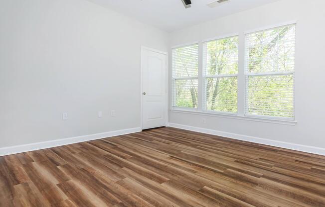 Empty room with large windows allowing natural light, featuring wooden flooring and a light-colored wall. A door is visible on the side, and no furniture is present, creating a spacious and airy atmosphere.