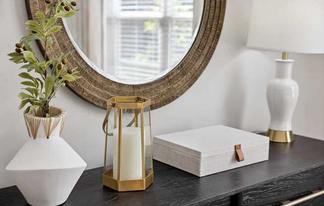 A white vase with a plant in it sits on a table next to a mirror and a white lamp.