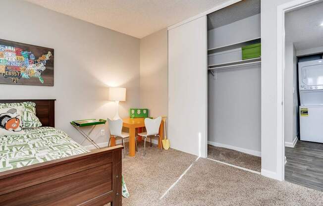 A bedroom with a bed, a table and chairs, and a refrigerator in the corner.