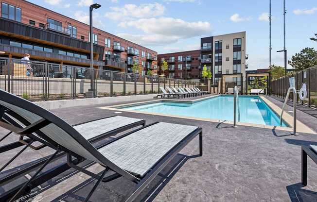 A pool area with sun loungers and a fence in front of apartment buildings.