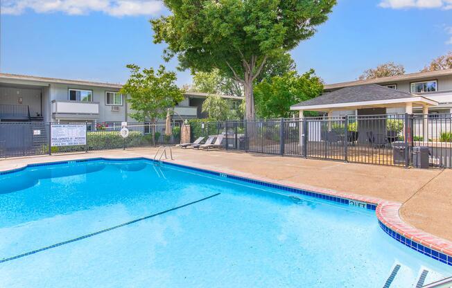 A clear blue swimming pool surrounded by a concrete deck. Lounge chairs are positioned nearby, and a fenced area features a shaded gazebo. Lush green trees and landscaping are visible in the background, with apartment buildings in the vicinity. Sunny outdoor setting ideal for relaxation.