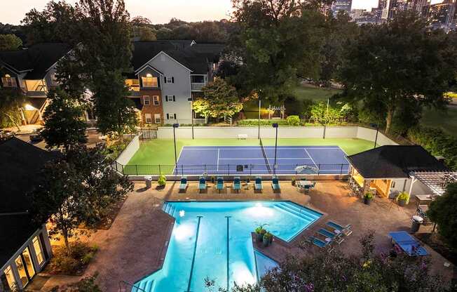 A tennis court is lit up at night with a pool in the foreground.