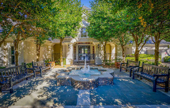 A tranquil courtyard at Saxony at Chase Oaks Apartments in Dallas, TX, featuring a central fountain surrounded by benches, lush green trees, and a shaded pathway leading to the entrance of the clubhouse.