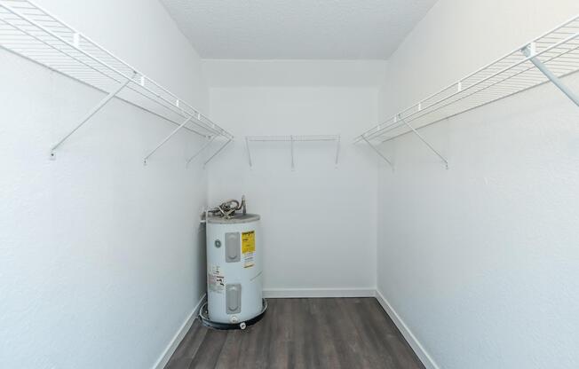 Empty storage room with white walls and a wooden floor. On the left side, there is a water heater with a yellow label, and several wire shelves mounted on the walls, creating space for storage. The overall space appears clean and well-lit.
