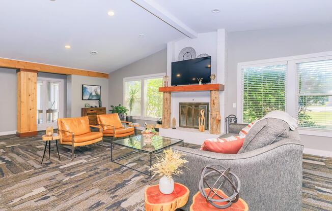 A modern living room with large windows letting in natural light. It features a gray sofa, two orange armchairs, a glass coffee table, and a stylish fireplace with a mounted TV above it. Decorative elements include a wooden beam and small plants, creating a cozy and inviting atmosphere.