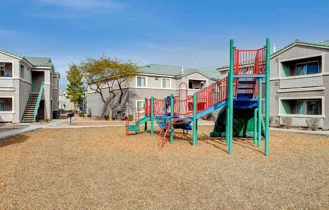 A playground with a red and blue slide in front of apartment buildings.