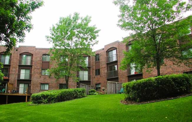 A grassy area in front of a brick building with trees and bushes.