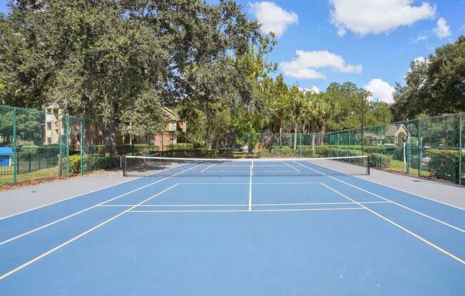 A blue tennis court surrounded by green fencing and trees.