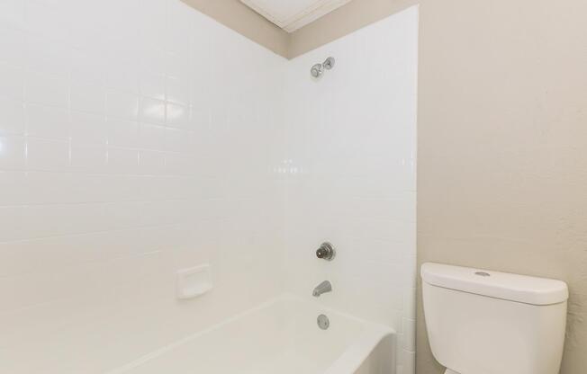 A clean and minimalist bathroom featuring a white tiled wall, a bathtub with a chrome faucet and showerhead, and a white toilet. The walls are a light beige, providing a neutral backdrop.
