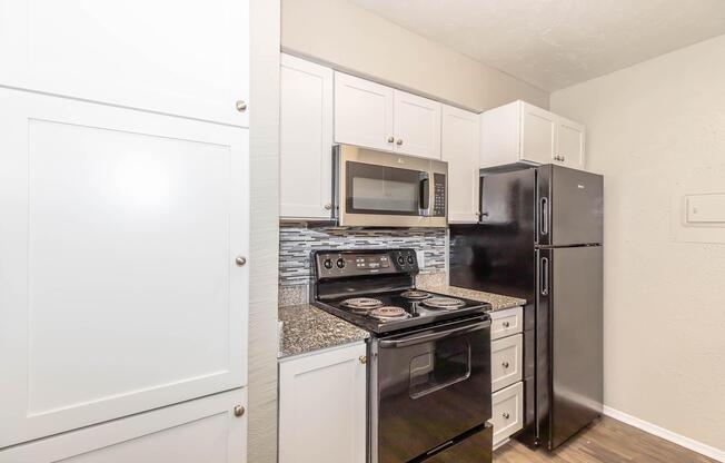 A modern kitchen featuring white cabinets, a stainless steel oven and microwave, and a black refrigerator. The countertop is made of granite, and the backsplash includes decorative tiles. The flooring is a dark wood laminate, contributing to the sleek design of the space.