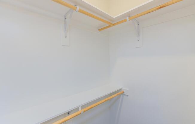 A clean, empty closet featuring white walls and a built-in shelf. Two wooden rods are mounted on the wall for hanging clothes, and the overall design is minimalistic and spacious, suitable for organizing garments and accessories.