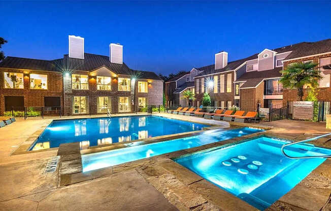 A resort-style pool area here at Lakewood on the Trail at night with a lit swimming pool, spa section, lounge chairs, and brick buildings, creating a vibrant, relaxing atmosphere.