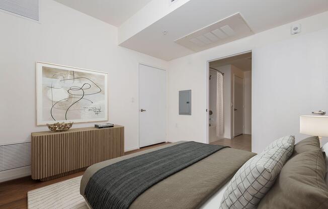 A modern bedroom featuring a neatly made bed with a grey and white bedspread, a decorative pillow, and a wooden dresser with a decorative bowl on top. A large framed abstract artwork is hung on the wall, and a door leads to a bathroom. Light-colored walls and a hardwood floor create a bright and open atmosphere.