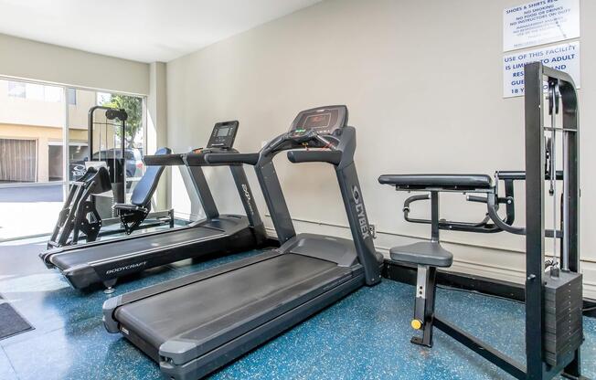 A small gym area featuring two treadmills and a weight bench. The equipment is neatly arranged on a blue floor, with large windows allowing natural light into the space. A sign on the wall provides facility guidelines and usage instructions.
