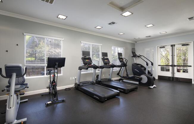 A brightly lit gym with modern fitness equipment, including treadmills, an elliptical machine, and a stationary bike. Large windows allow natural light to fill the space, and there are doors leading outside. The floor is dark, and the walls are painted light gray, creating a clean and inviting atmosphere for workouts.
