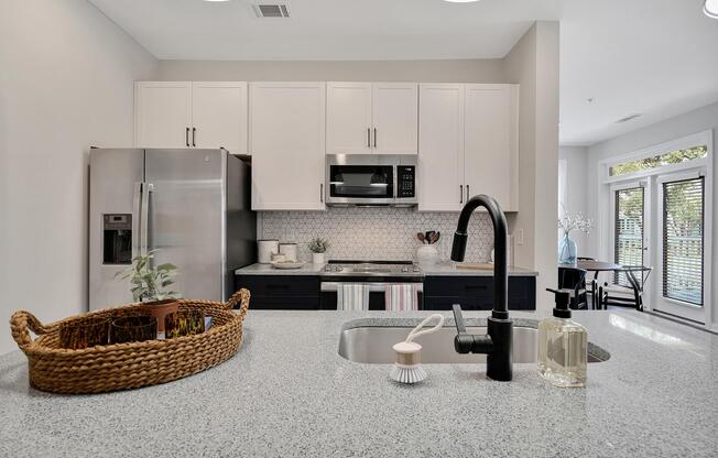 A modern kitchen featuring a stainless steel refrigerator and microwave, white cabinetry, and a granite countertop. A woven basket holds plants and kitchen items, while a sleek black faucet is visible. The background includes a dining area with large windows and a table.