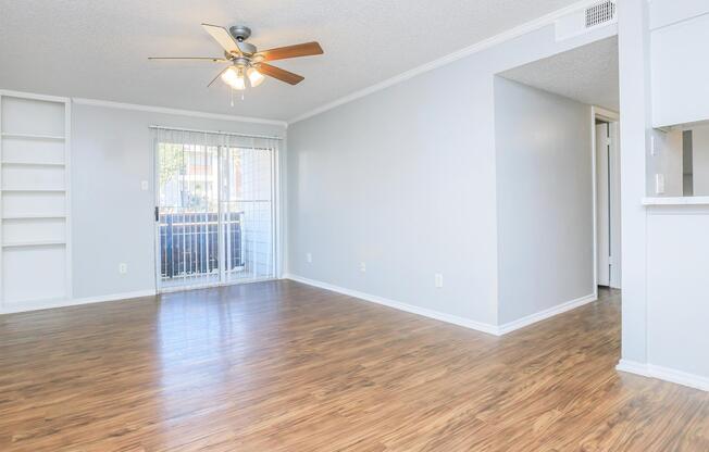 Spacious interior featuring light-colored walls and a ceiling fan. The room has a modern laminate floor, a large window with sliding glass doors leading to a balcony, and built-in shelves on one wall. A doorway to another room is visible on the right side, providing a bright and airy atmosphere.