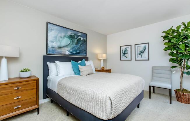 A cozy bedroom featuring a comfortable bed with light bedding and decorative pillows. There are bedside tables with lamps on either side, a chair, and framed artwork depicting ocean waves and botanical prints on the walls. A potted plant adds a touch of greenery to the space.