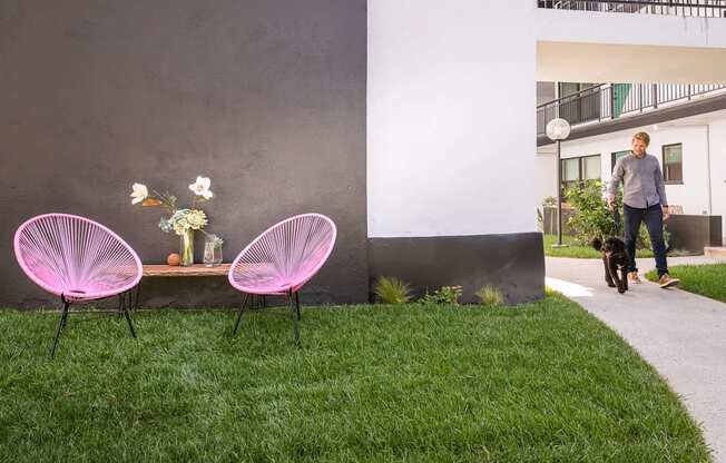 A man walking his dog next to a pink chair and table with flowers on it.