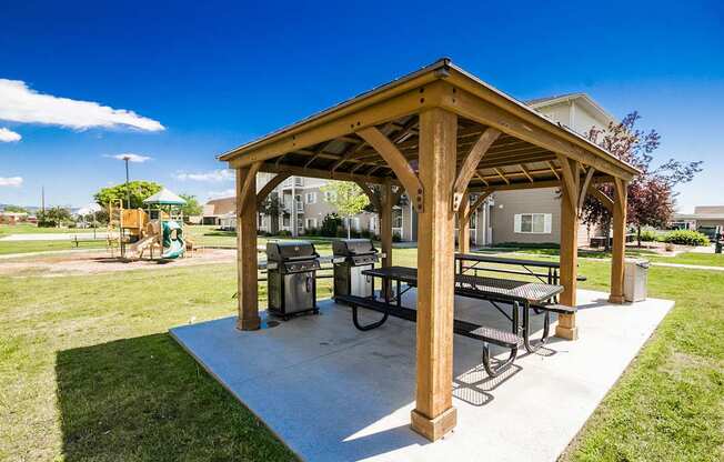 A wooden pavilion with a picnic table and chairs is situated in a grassy area.