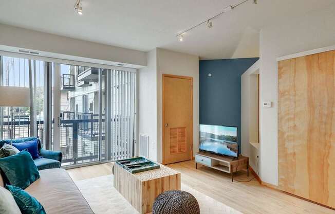 A modern living room here at Blue and Lime showcasing light wood flooring, a plush sofa with blue accent pillows, and a low-profile TV console set against a bold blue accent wall. Large sliding glass doors with vertical blinds open to a private balcony, allowing natural light to fill the space and highlight the clean, contemporary finishes.