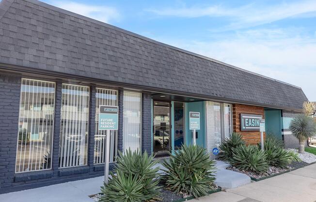 A modern building with a flat roof and large windows. The exterior features a mix of dark bricks and wood accents. In front, there are neatly arranged succulents and a sign indicating "Future Resident Parking." The overall appearance is clean and inviting, set against a blue sky.