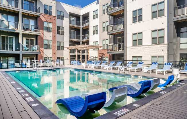 A pool with blue lounge chairs and a wooden deck in front of apartment buildings.