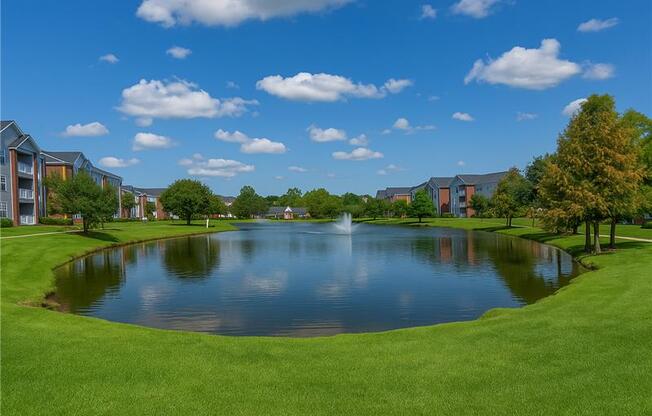 A serene landscape featuring a pond, a fountain, and a row of buildings.