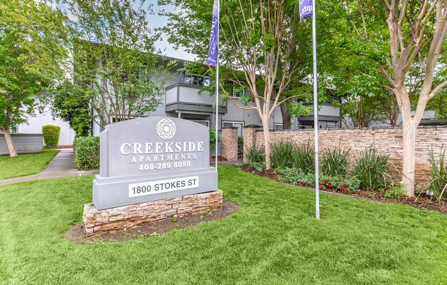 Sign for Creekside Apartments located at 1800 Stokes St, featuring a landscaped entrance with greenery and decorative stonework. Two flags are displayed nearby.