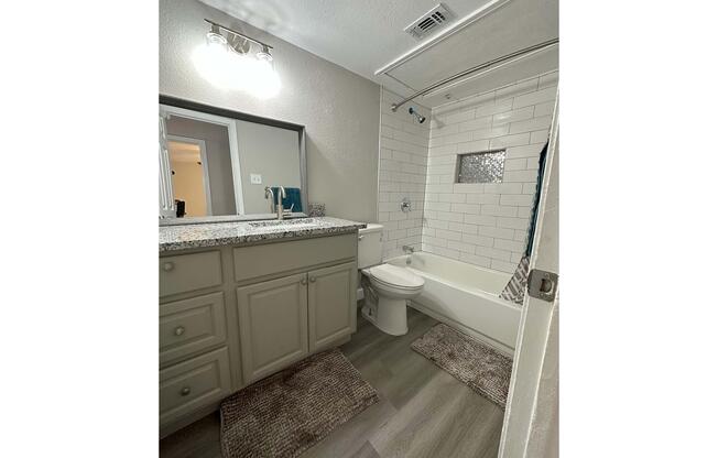 A clean and modern bathroom featuring a bathtub with a shower, a granite countertop vanity with a mirror, and light fixtures. The floor is covered with light-colored vinyl planks, and plush area rugs are placed near the tub and sink. The walls have white subway tiles and soft neutral paint.