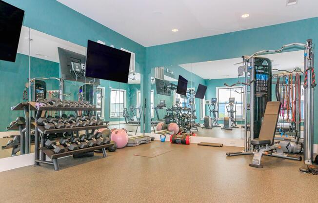 A spacious gym area featuring various workout equipment. There are racks of dumbbells, exercise balls, and a multi-station strength training machine. Large mirrors reflect the equipment, and two flat-screen TVs are mounted on the wall, providing a modern fitness environment. The flooring is cushioned for comfort.