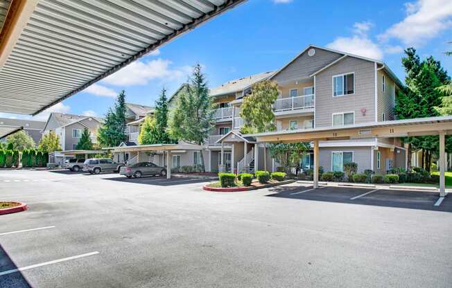A covered parking area here at Allegro showcasing reserved carport spaces in front of three-story apartment buildings with neutral siding, private balconies, and exterior staircases, surrounded by landscaped greenery, mature trees, and paved drive lanes under a bright blue sky.
