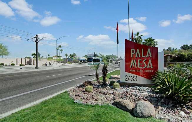 A vibrant community monument sign here at Pala Mesa featuring bold red lettering and modern design, set against manicured green grass and desert landscaping with palm trees and decorative stone accents. Positioned along a main roadway with nearby transit access, the welcoming entrance highlights the property’s convenient location and curb appeal.